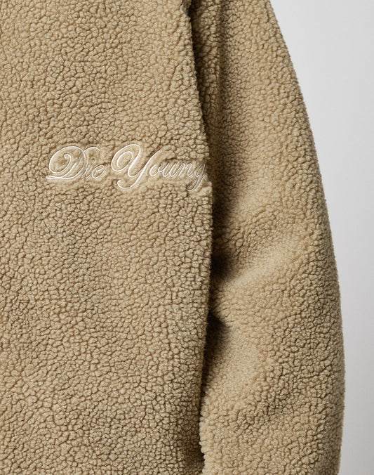 Close-up of the LFDY Teddy Jacket by LIVE FAST DIE YOUNG—a beige, fluffy, oversized jacket with a zipper and “Die Young” embroidered in white cursive on the chest.