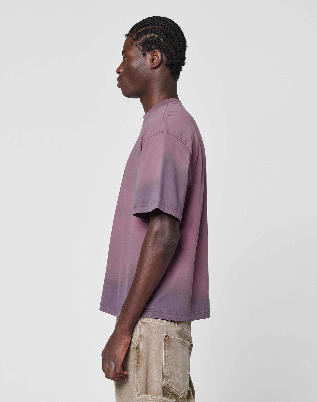 A person with braided hair in profile wears the LIVE FAST DIE YOUNG Live Fast District Tee, a loose-fitting, short-sleeve purple ombré t-shirt with Spray Effekt, paired with beige pants against a plain white background.