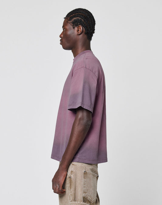 A person with braided hair in profile wears the LIVE FAST DIE YOUNG Live Fast District Tee, a loose-fitting, short-sleeve purple ombré t-shirt with Spray Effekt, paired with beige pants against a plain white background.