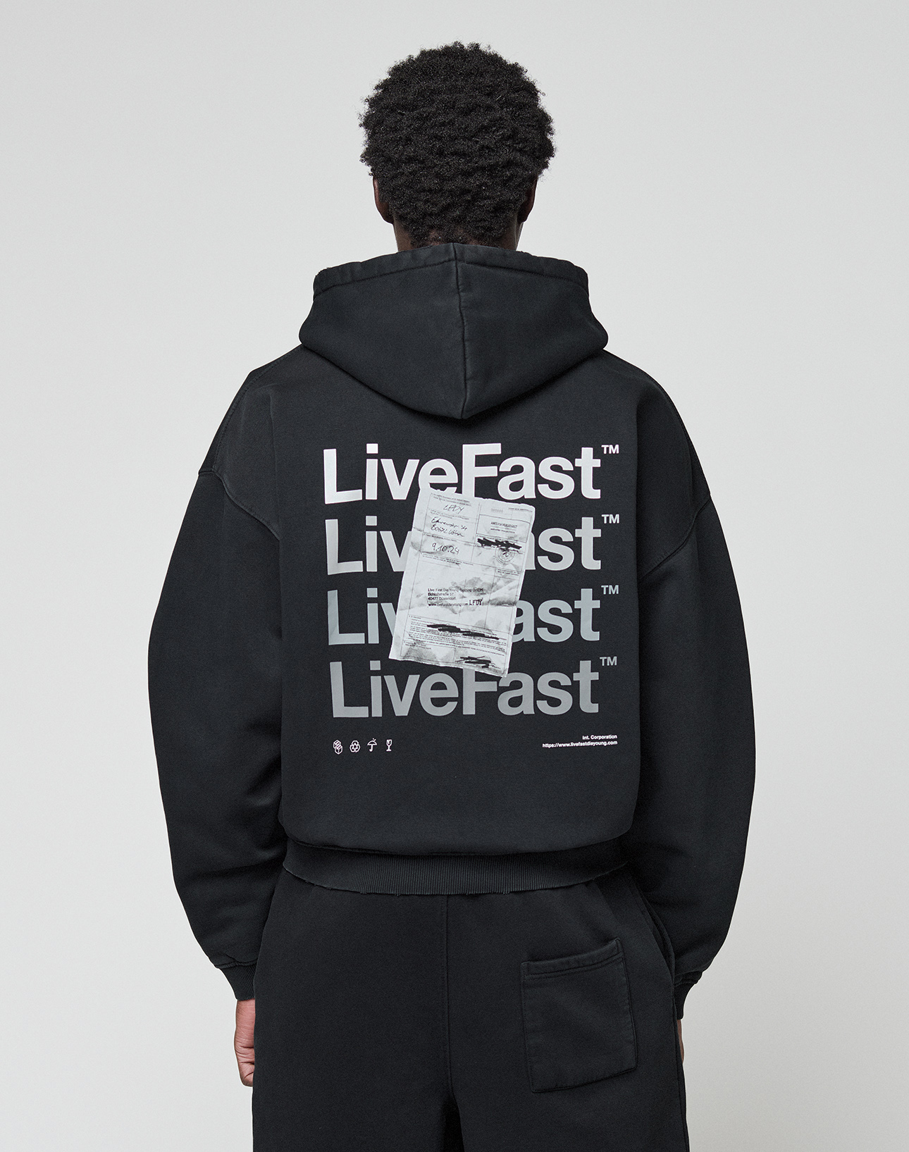 A person wearing the LIVE FAST DIE YOUNG Basic Evidence Hooded Zip in an oversize fit, featuring bold white branding, paper-like graphic, small symbols, and subtle distressing on the back, stands against a plain background. | model