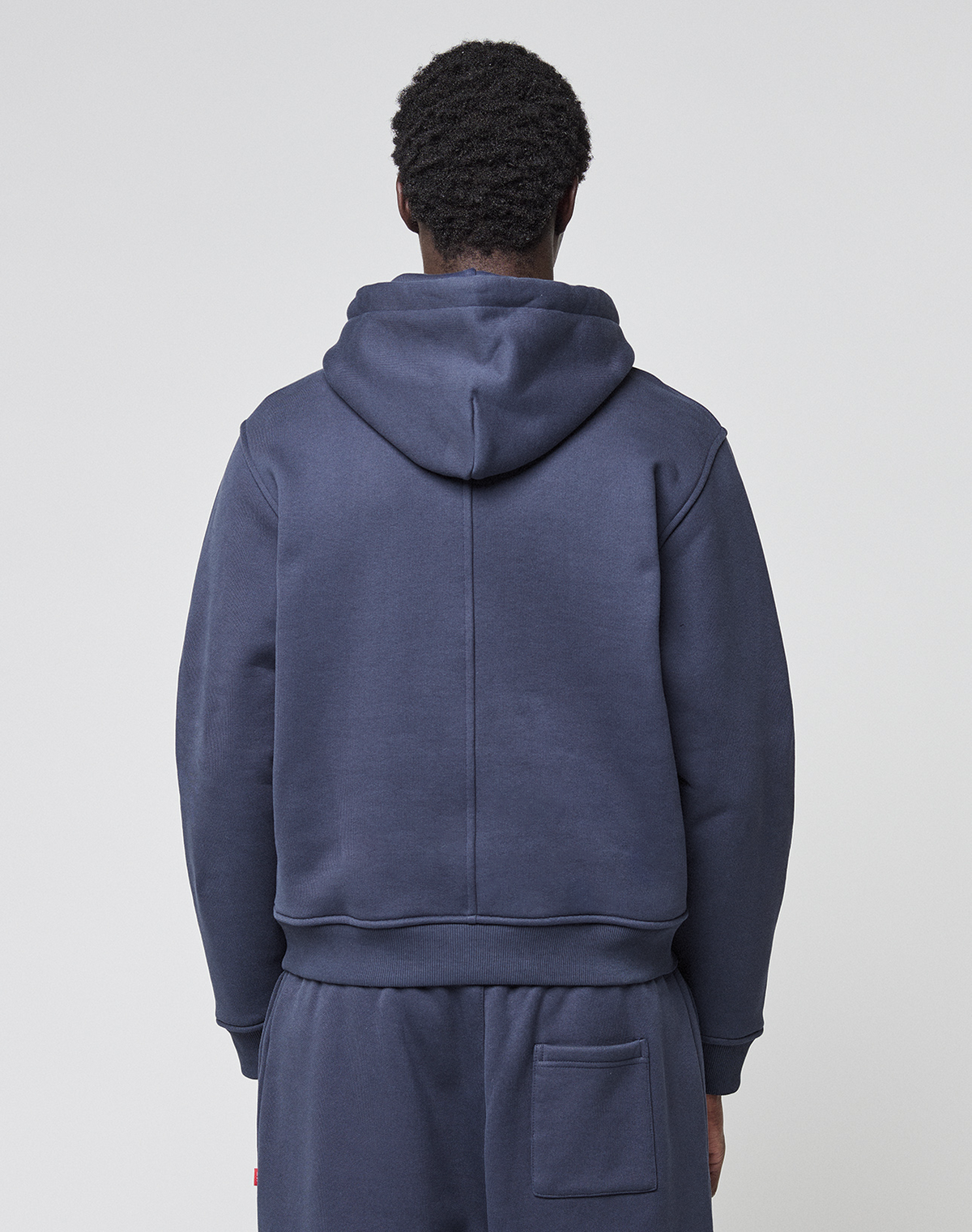 A person with short curly hair is seen from behind, wearing the LIVE FAST DIE YOUNG LFDY Heavyweight Lounge Hooded in navy blue with matching pants featuring a back pocket, against a plain light background. Made from 100% Baumwolle.