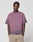 A man with braided hair wears the LIVE FAST DIE YOUNG Live Fast District Tee, an oversized faded purple T-shirt with a subtle spray effect, paired with beige pants, standing hands-in-pockets against a plain white background.