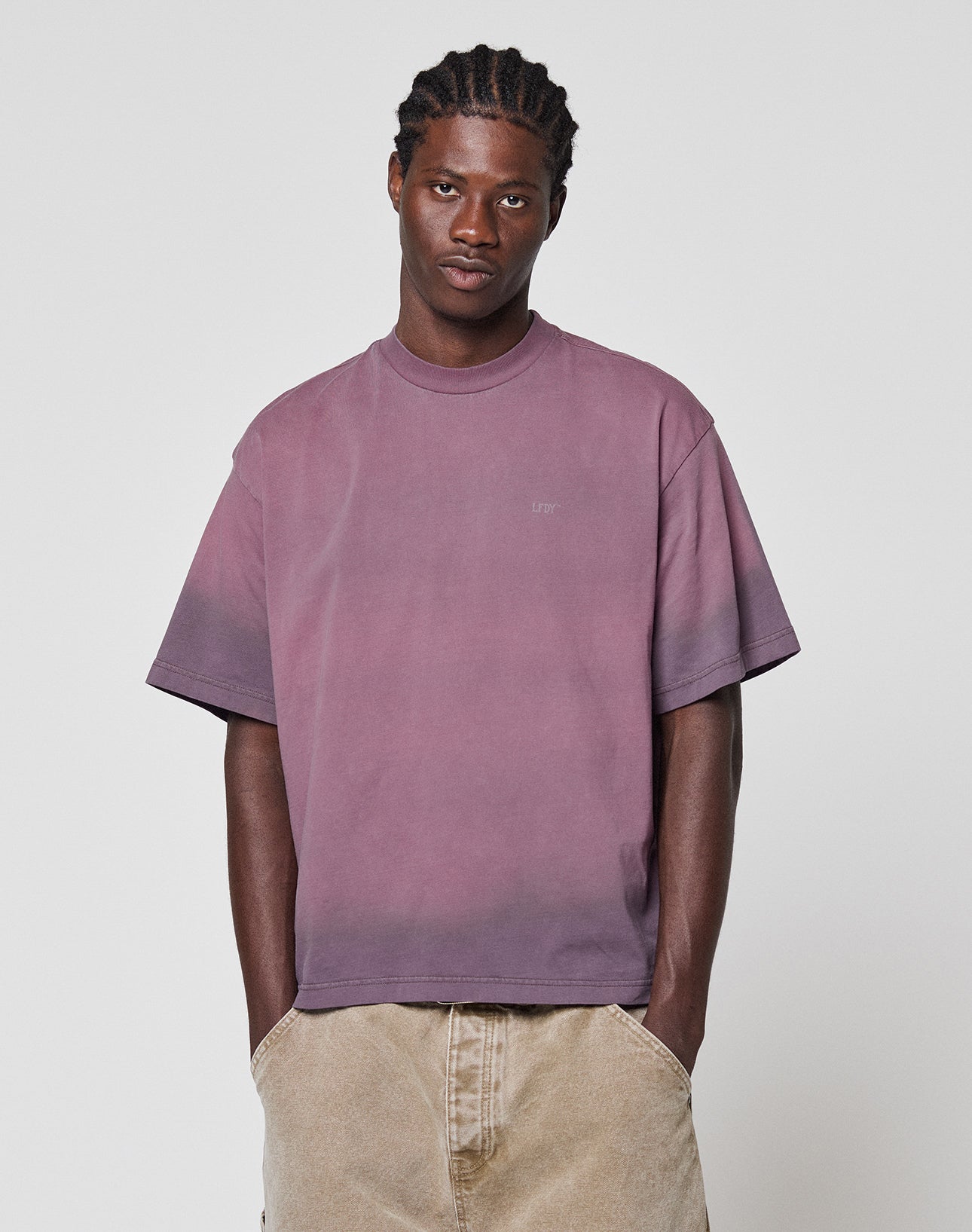 A man with braided hair wears the LIVE FAST DIE YOUNG Live Fast District Tee, an oversized faded purple T-shirt with a subtle spray effect, paired with beige pants, standing hands-in-pockets against a plain white background.