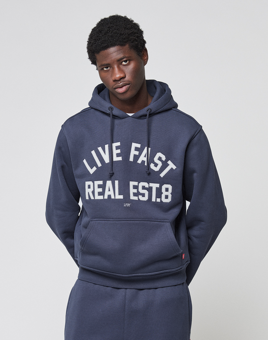 A young man wearing the LIVE FAST DIE YOUNG LFDY Heavyweight Lounge Hooded in dark blue and matching pants stands against a plain background. The 100% Baumwolle hoodie features "LIVE FAST REAL EST.8" in white on the front.