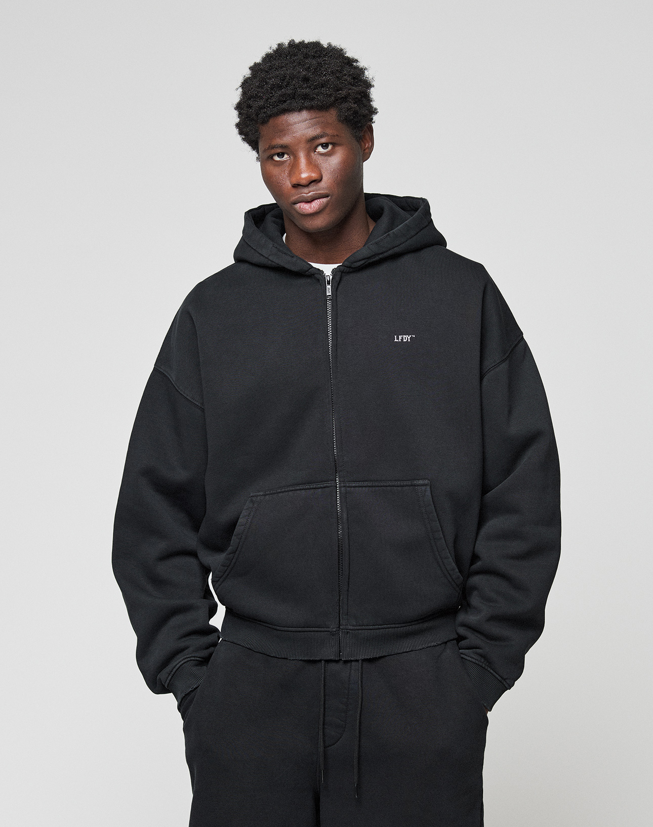A person wears the Basic Evidence Hooded Zip by LIVE FAST DIE YOUNG, featuring subtle distressing and branding, paired with oversized black pants. They stand against a light gray background, looking at the camera with hands in their pockets.