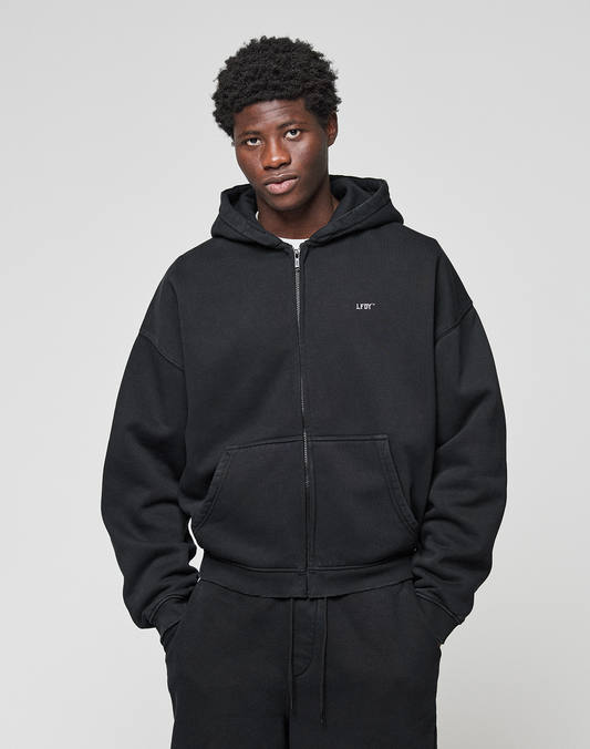 A person wears the Basic Evidence Hooded Zip by LIVE FAST DIE YOUNG, featuring subtle distressing and branding, paired with oversized black pants. They stand against a light gray background, looking at the camera with hands in their pockets.