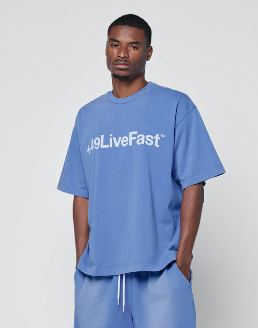 A man stands with his hands in his pockets against a light gray background, wearing the LIVE FAST DIE YOUNG Basic +49 Tee—an oversize blue T-shirt made from weicher Baumwolle with “+49LiveFast” in white—and matching shorts.