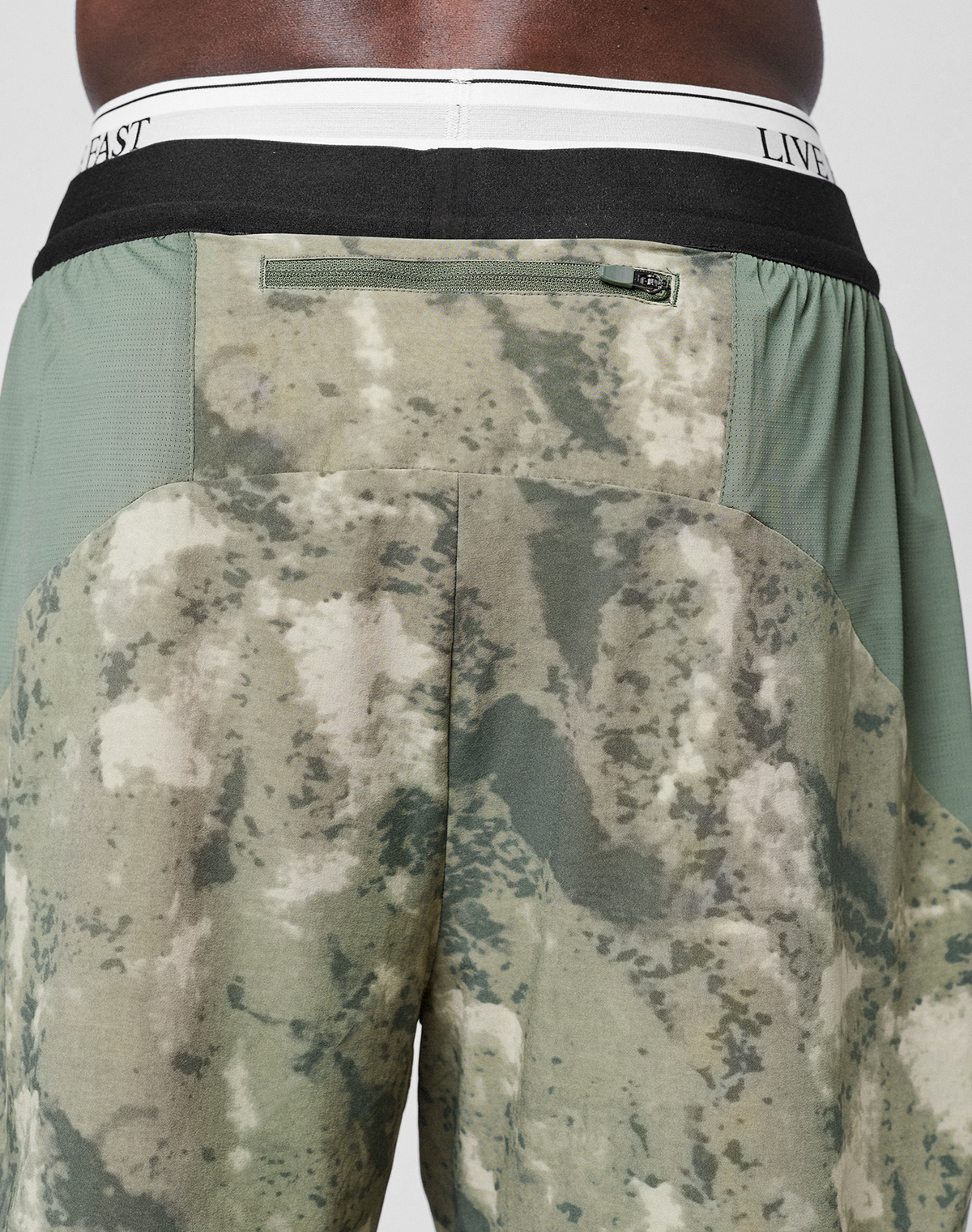 Close-up of the back of a person wearing green and tan camouflage LFDY Performance Running Shorts by LIVE FAST DIE YOUNG, featuring a black waistband, white elastic band, and reflective LIVEFAST logo print.