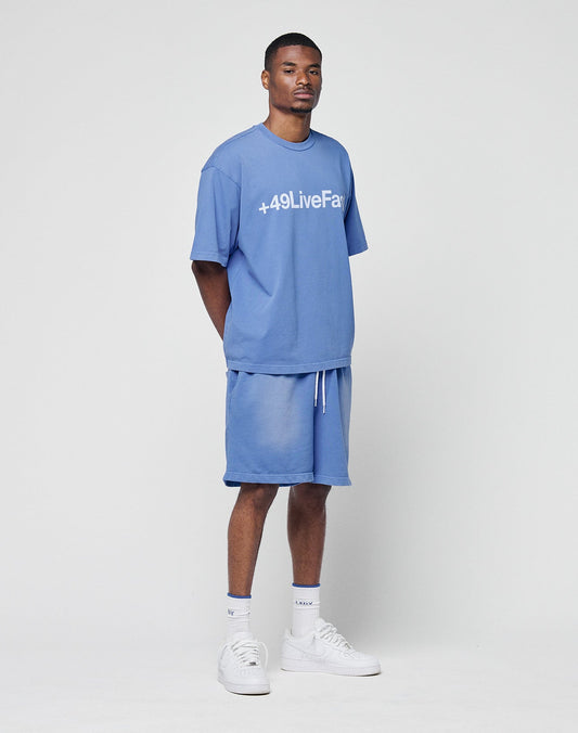 A man stands against a white background wearing the Basic +49 Tee by LIVE FAST DIE YOUNG in light blue, with +49LiveFair printed on the front, matching blue shorts, white socks, and white sneakers. His arms are behind his back.