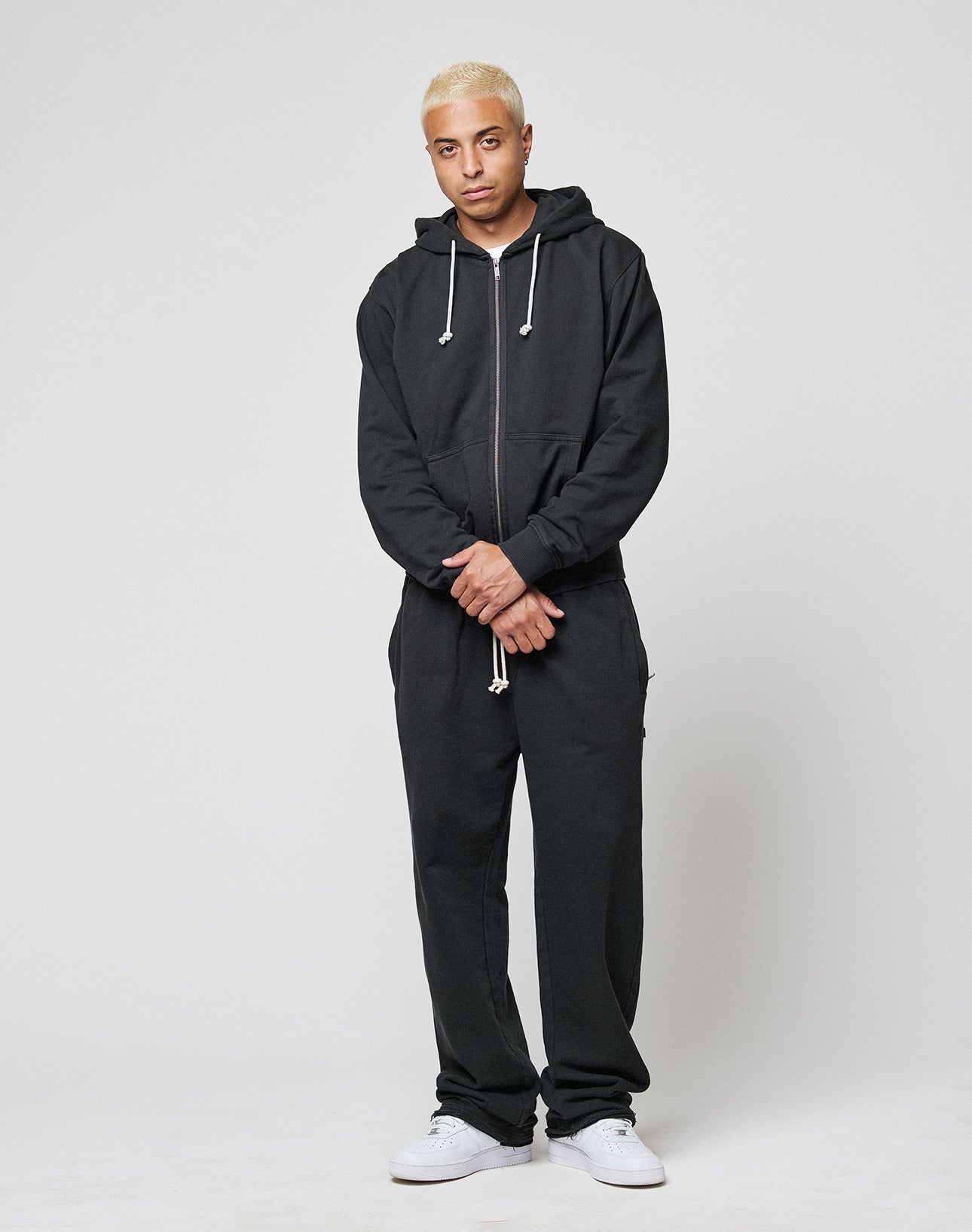 A person with short, bleached blond hair stands against a plain background, wearing the LIVE FAST DIE YOUNG LFDY Lightweight Jogger in an oversize fit and a black zip-up hoodie, paired with white sneakers. Their hands are loosely clasped in front.