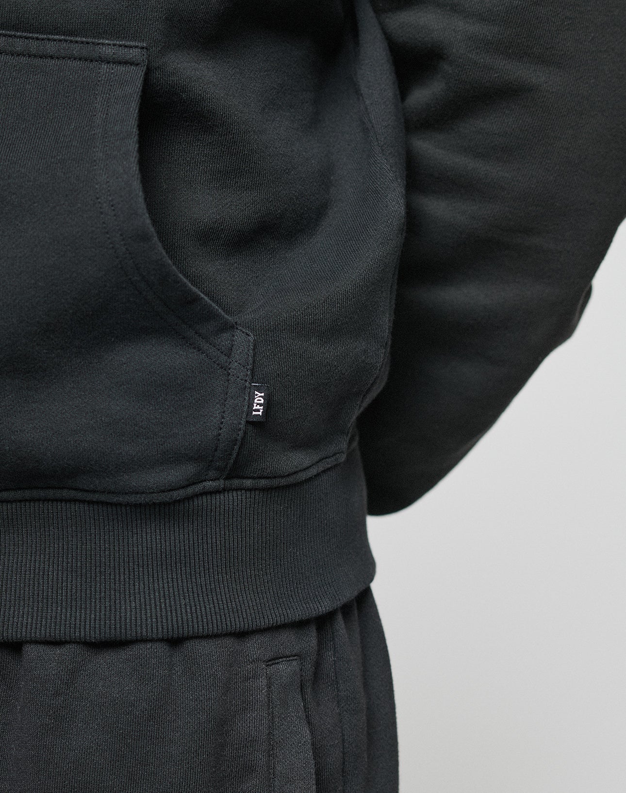 Close-up of a person in a black LFDY Lightweight Hooded Zip by LIVE FAST DIE YOUNG, paired with matching pants. The lower side of the hoodie features a small black tag with white text. Made from 100% Baumwolle. The hand is not visible.