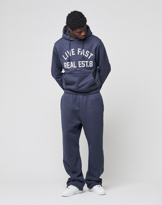 Wearing the LIVE FAST DIE YOUNG LFDY Heavyweight Lounge Hooded and matching sweatpants, a person in a dark blue 100% Baumwolle set with "LIVE FAST REAL EST.8" stands against a light background, hands behind their back. Regular Fit offers comfort and style.