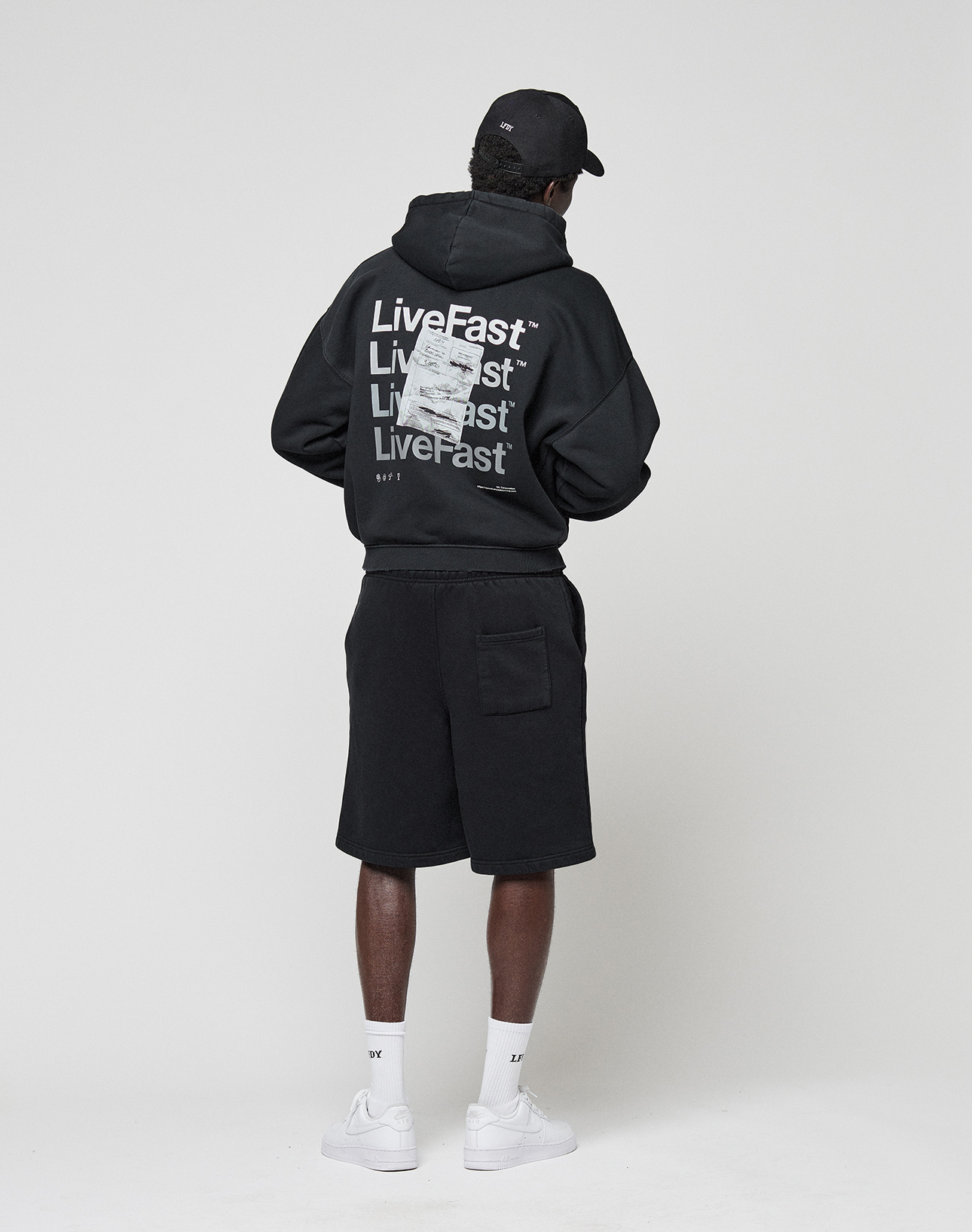 A person wears the LIVE FAST DIE YOUNG Basic Evidence Hooded Zip in black, paired with black shorts, white socks, white sneakers, and a black cap, standing with their back to a plain light background.
