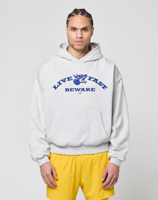 A person with braided hair wears the LIVE FAST DIE YOUNG Live Fast Beware Hooded, a light gray oversized double-layer hoodie with a bold front print, styled with blue and yellow shorts against a plain light background. | model