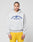 A person with braided hair wears the LIVE FAST DIE YOUNG Live Fast Beware Hooded, a light gray oversized double-layer hoodie with a bold front print, styled with blue and yellow shorts against a plain light background.