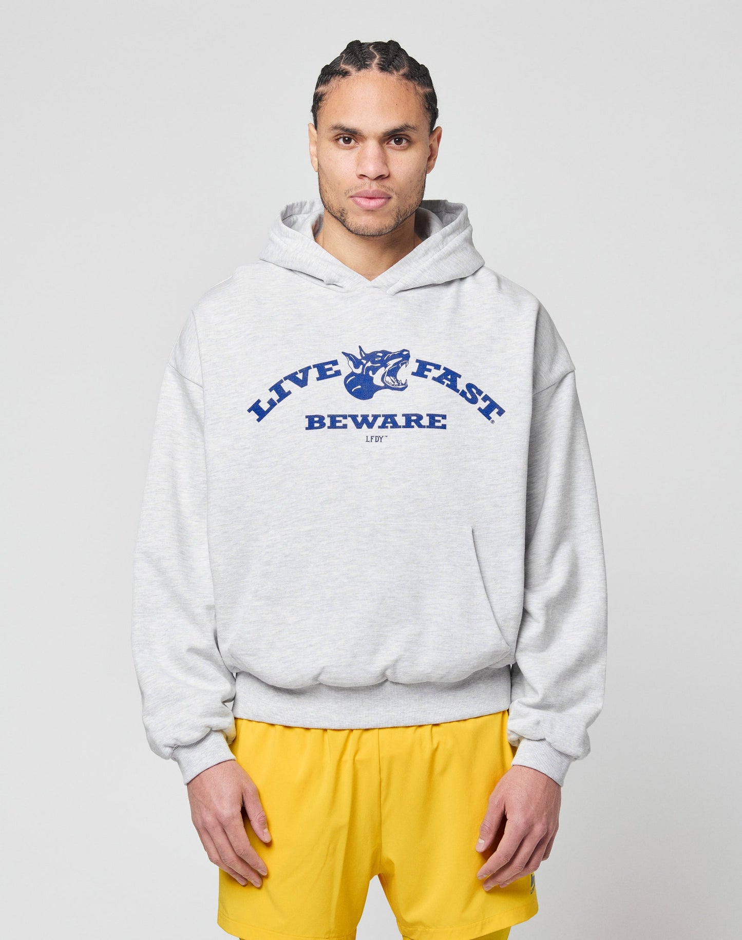 A person with braided hair wears the LIVE FAST DIE YOUNG Live Fast Beware Hooded, a light gray oversized double-layer hoodie with a bold front print, styled with blue and yellow shorts against a plain light background. | model
