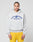 A person with braided hair wears the LIVE FAST DIE YOUNG Live Fast Beware Hooded, a light gray oversized double-layer hoodie with a bold front print, styled with blue and yellow shorts against a plain light background.