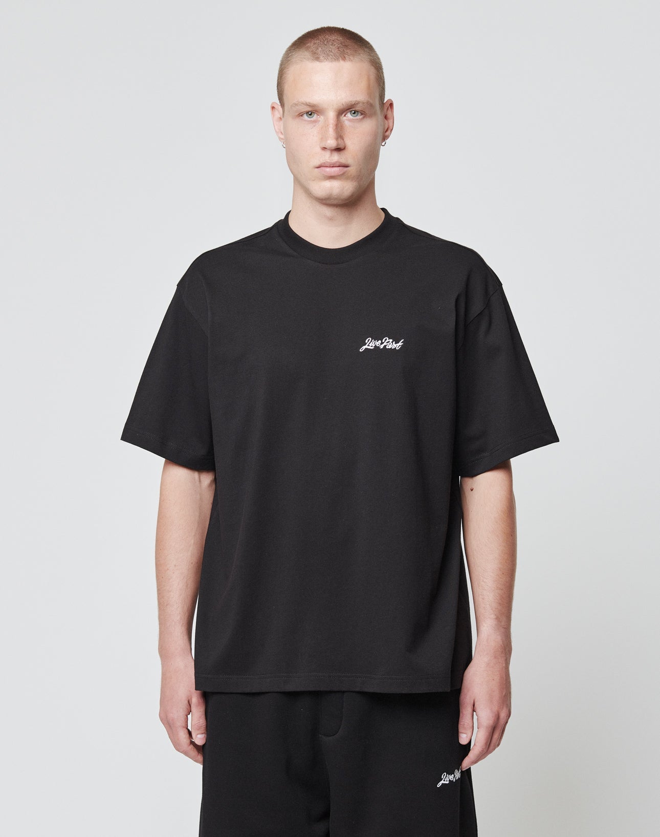 A person with short hair wears the LIVE FAST DIE YOUNG Basic Script Tee in loose black Oversize Fit and black pants, standing against a plain light gray background. The tee features a small white logo embroidery on the left chest.
