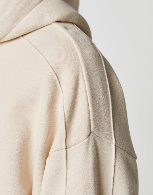 Close-up of the shoulder and upper sleeve of the cream-colored LFDY Pleated Hooded Zip by LIVE FAST DIE YOUNG, showing cotton fabric texture and seam details. The drawstring hood is partially visible in the upper left corner.
