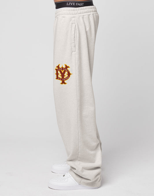 A person wears DY Jogger sweatpants by LIVE FAST DIE YOUNG in light gray with wide legs, oversize fit, black waistband featuring LIVE FAST text, yellow and red embroidered crest on the side, white sneakers, and a plain white background.