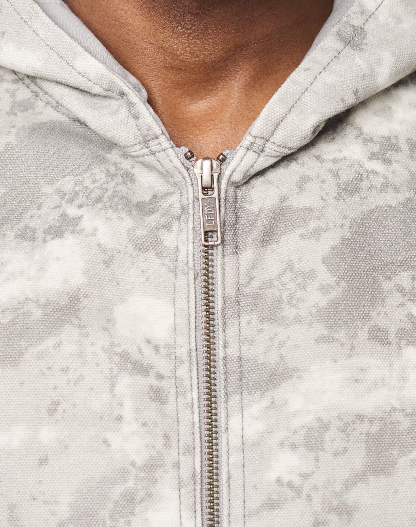Close-up of a person wearing the Live Fast Canvas Jacket by LIVE FAST DIE YOUNG, featuring a light gray, textured marbled pattern, metal zipper pulled up most of the way, and an oversize fit showing the upper chest and neck.