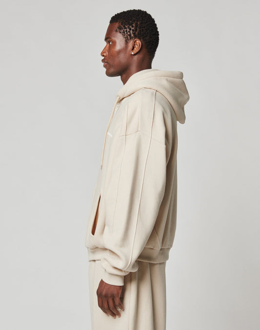 A man in profile wears the LIVE FAST DIE YOUNG LFDY Pleated Hooded Zip in an oversized beige fit with matching sweatpants against a plain, light background.