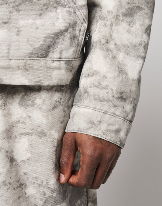 A close-up of a right hand resting by the side, wearing the Live Fast Canvas Jacket from LIVE FAST DIE YOUNG in light grey/off-white tie-dye. The focus is on the textured fabric and sleeves black tag.
