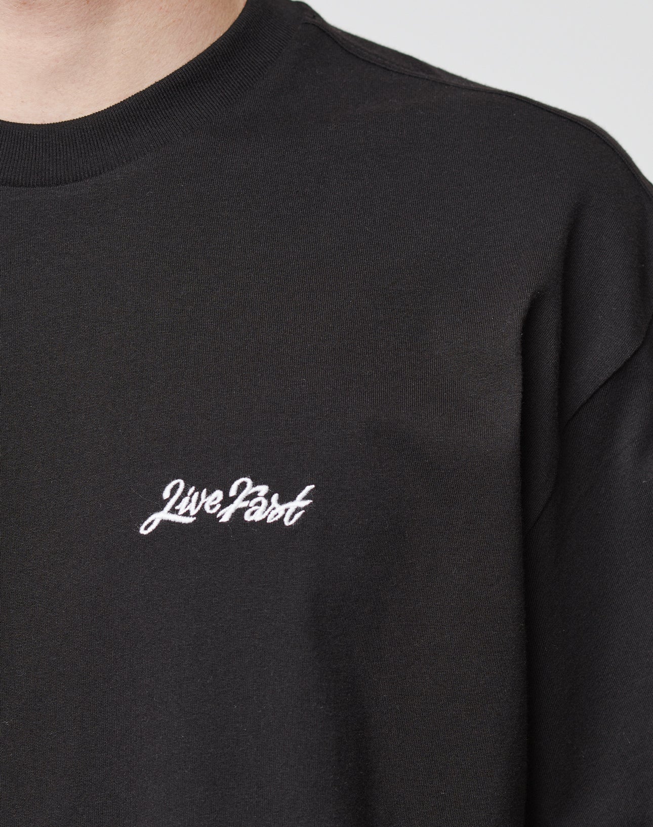 Close-up of a person wearing a black Basic Script Tee by LIVE FAST DIE YOUNG, with Live Fast in white script on the left chest. Only the neck, shoulder, and upper torso are shown. Made from 100% cotton in an oversize fit.