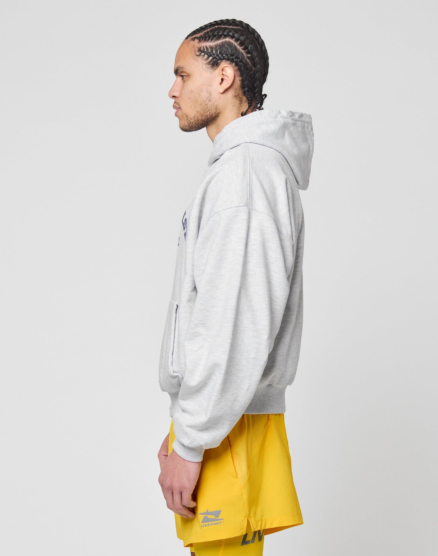 A person with braided hair, wearing the LIVE FAST DIE YOUNG Live Fast Beware Hooded in light gray and bright yellow shorts, stands in profile against a plain white background.