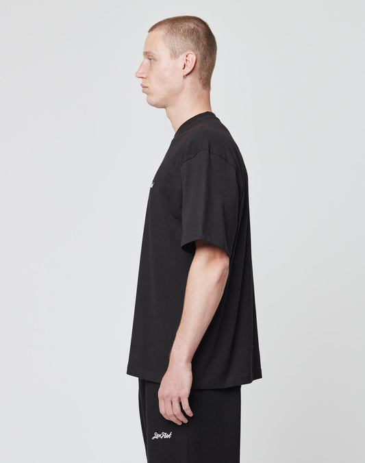 A man with short hair stands in profile against a plain background, wearing a LIVE FAST DIE YOUNG Basic Script Tee in black and matching black pants with white text by the pocket, both made from soft Baumwolle.