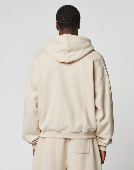 A person in an oversize fit, light beige LFDY Pleated Hooded Zip by LIVE FAST DIE YOUNG with Kapuze mit Kordel and matching sweatpants, made from soft Baumwolle, stands facing away against a plain light gray background.