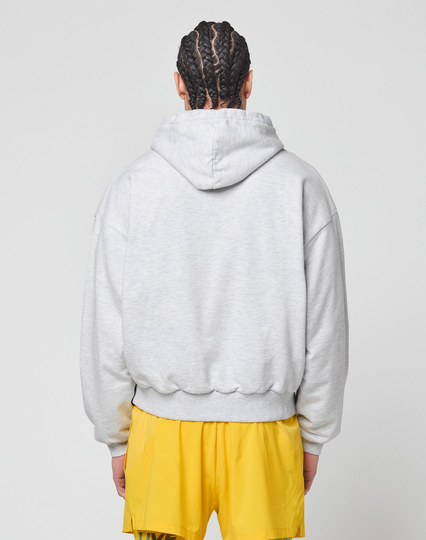 A person with braided hair is seen from the back, wearing the LIVE FAST DIE YOUNG Live Fast Beware Hooded in an oversize fit, paired with bright yellow shorts against a plain light background.