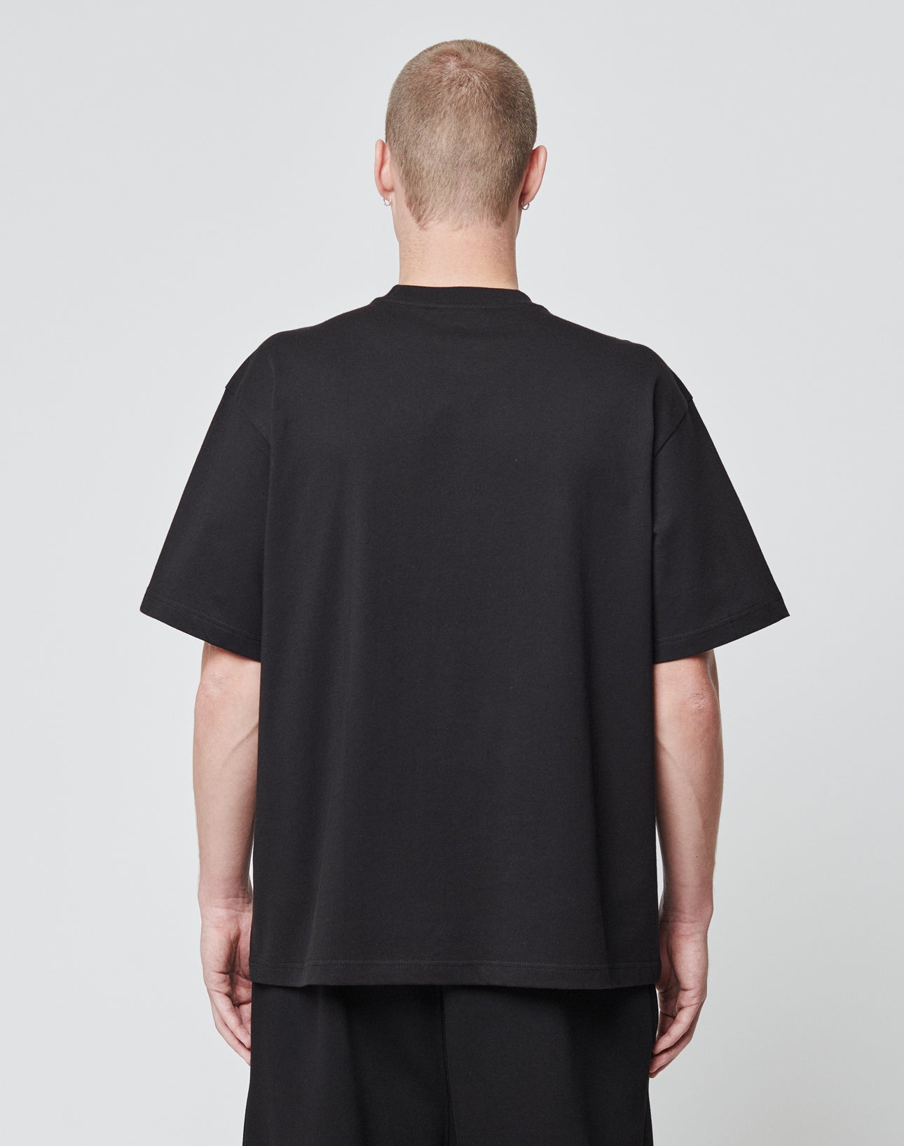 A person with short hair stands facing away from the camera, wearing an oversize fit LIVE FAST DIE YOUNG Basic Script Tee in black cotton and black shorts against a plain light gray background.