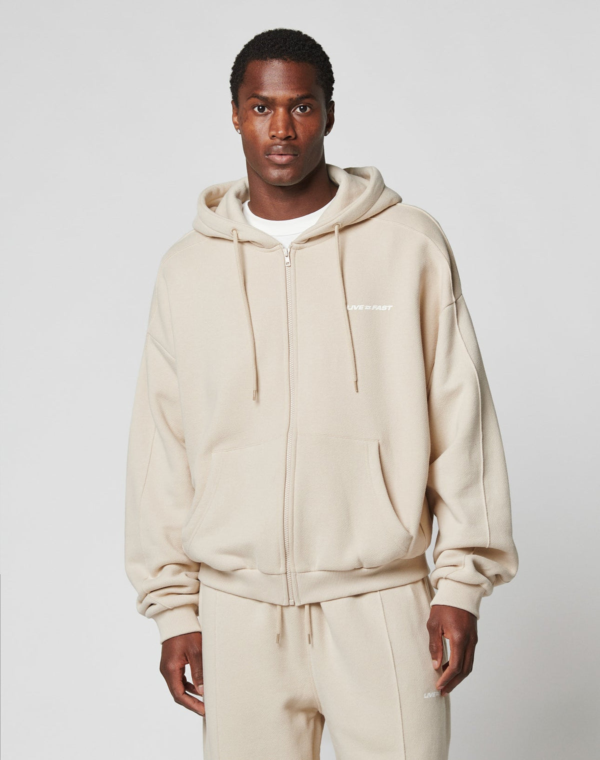 A man wearing an LFDY Pleated Hooded Zip by LIVE FAST DIE YOUNG and matching beige sweatpants, both made from cotton, stands facing forward against a plain light gray background.