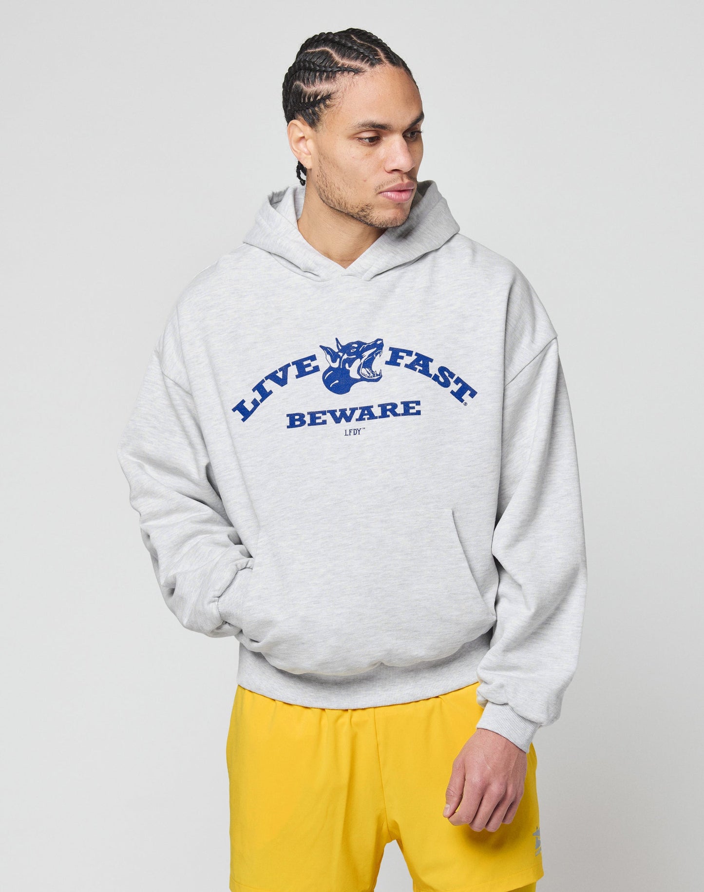 A man wearing the LIVE FAST DIE YOUNG Live Fast Beware Hooded with a front panther graphic, paired with yellow shorts, stands against a plain white background.