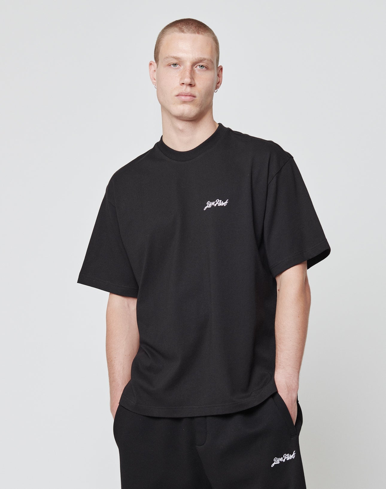 A person with short hair wears the LIVE FAST DIE YOUNG Basic Script Tee and matching black Baumwolle pants, both with small white Live Fast logo details, standing against a plain light background with hands in pockets.