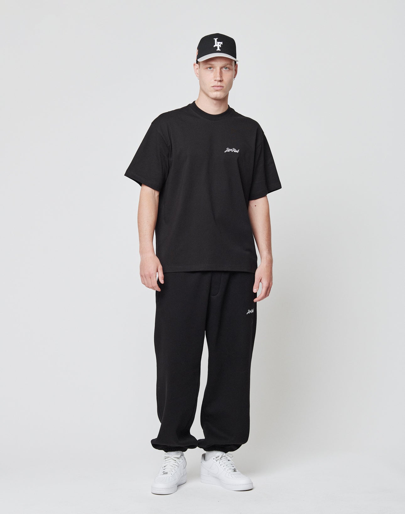 A man stands against a plain white background in a LIVE FAST DIE YOUNG Basic Script Tee, black sweatpants with logo embroidery, a matching cap, and white sneakers. Sporty logos accentuate his casual look throughout the outfit.