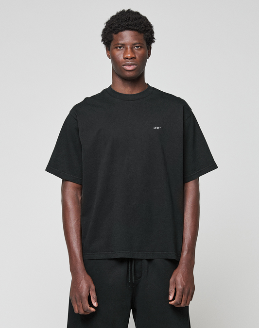 A young man in the LIVE FAST DIE YOUNG Basic Evidence Tee—an oversize fit black t-shirt made from 100% Baumwolle with short sleeves and small white chest text—stands against a light gray background, paired with black pants and a neutral expression.