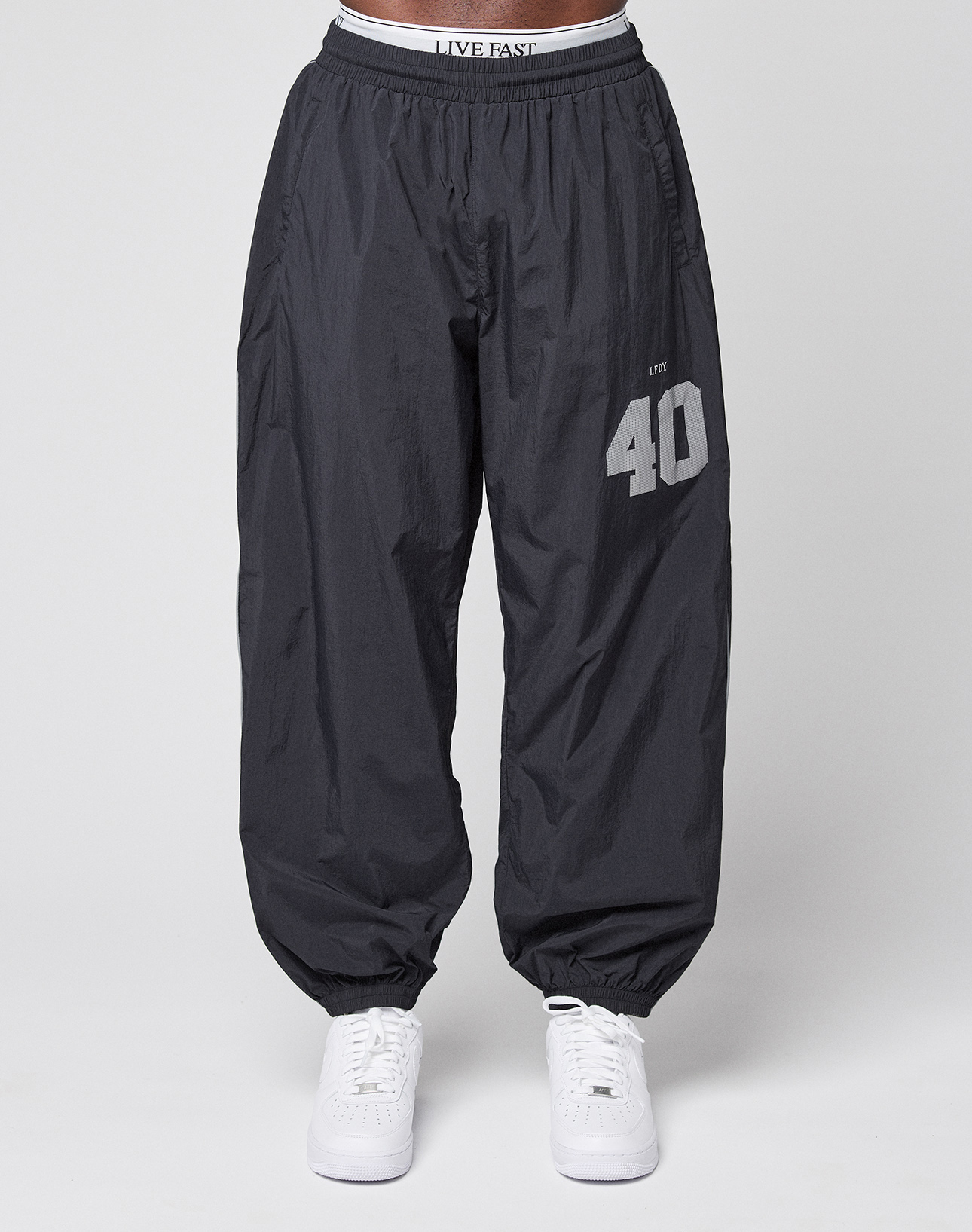 A person wears LFDY Performance Trackpants by LIVE FAST DIE YOUNG—oversized, 100% nylon, black with "40" and "LIVE" reflective print on the left thigh, elastic waistband and cuffs—paired with white sneakers on a plain light gray background.