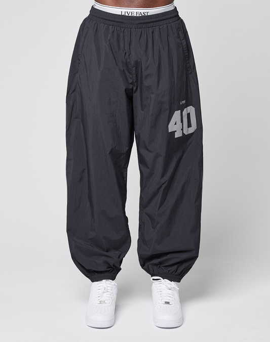 A person wears LFDY Performance Trackpants by LIVE FAST DIE YOUNG—oversized, 100% nylon, black with "40" and "LIVE" reflective print on the left thigh, elastic waistband and cuffs—paired with white sneakers on a plain light gray background.