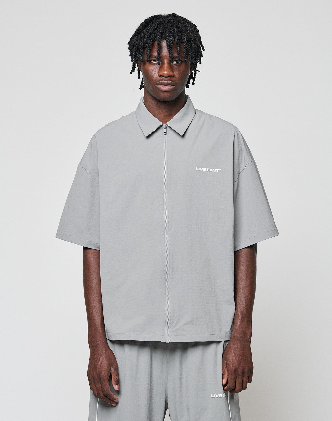 A man with short dreadlocks wears the LIVE FAST DIE YOUNG LFDY Ripstop Shirt and matching gray shorts, both with bold LIVE FAST Silikonprint text, standing against a plain light background.