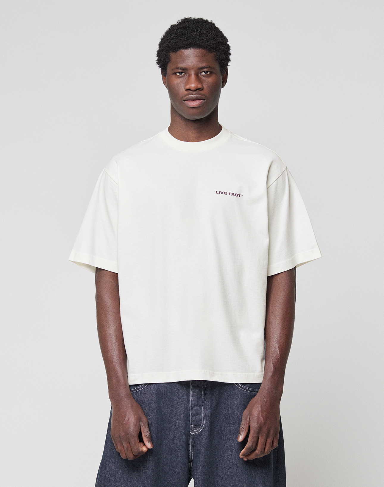 A man stands facing forward against a plain background, wearing the LIVE FAST DIE YOUNG LFDY Grand Theft Tee in white with front and back print, paired with dark blue jeans. The tee is made from 100% Baumwolle; his arms are relaxed at his sides.