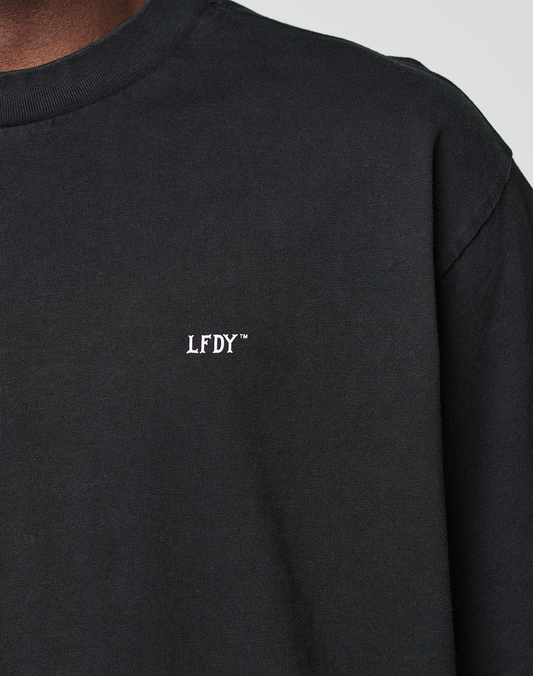A close-up of a person wearing the LIVE FAST DIE YOUNG Basic Evidence Tee, featuring a plain black oversized fit and small white “LFDY™” text embroidered on the chest, focusing on the upper torso and shoulder.
