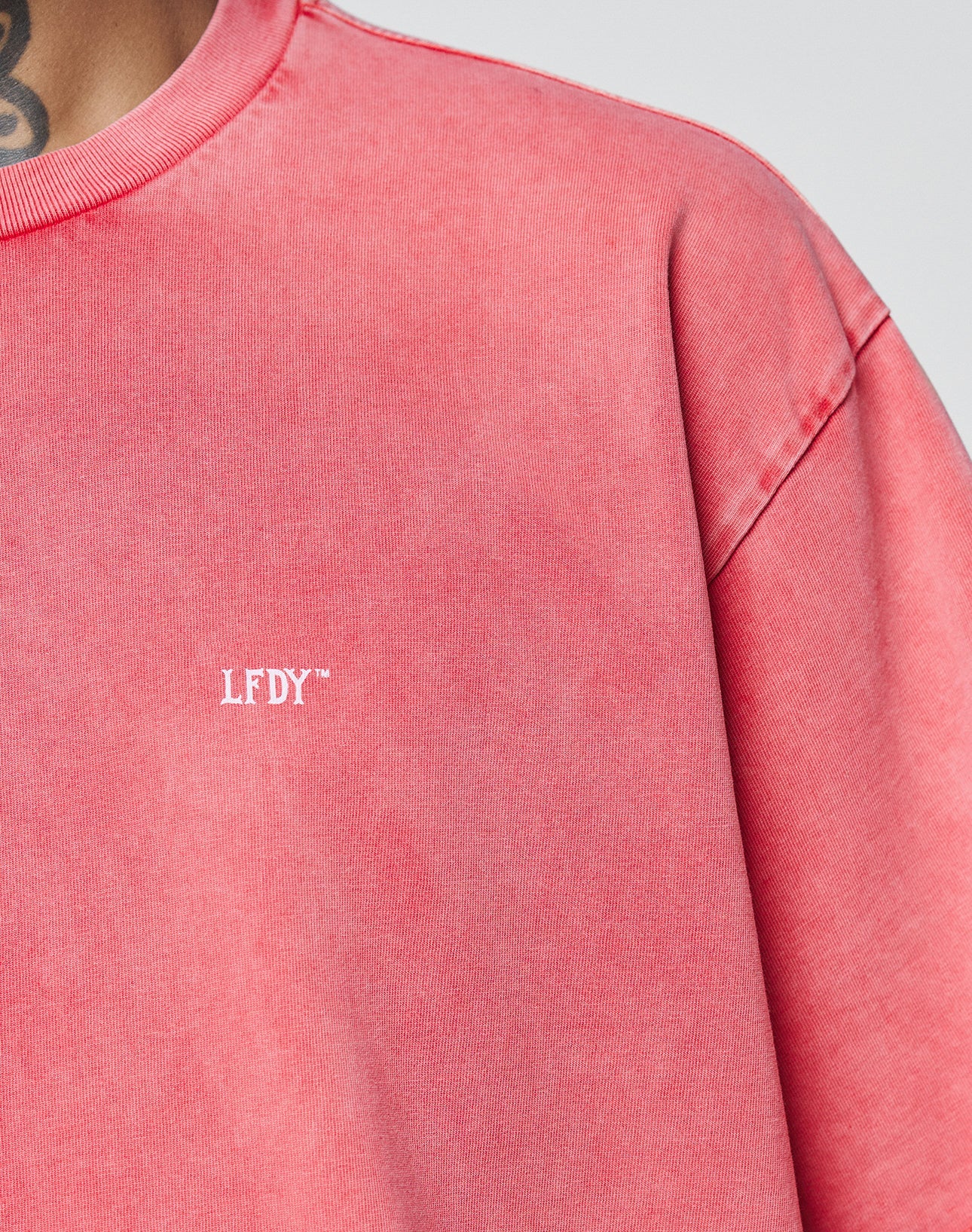 Close-up of a person wearing the LIVE FAST DIE YOUNG Basic Evidence Tee, an oversized faded pink shirt made from 100% Baumwolle, featuring small white LFDY™ text on the chest. Part of their neck, a tattoo, and the Flag-Label are visible.