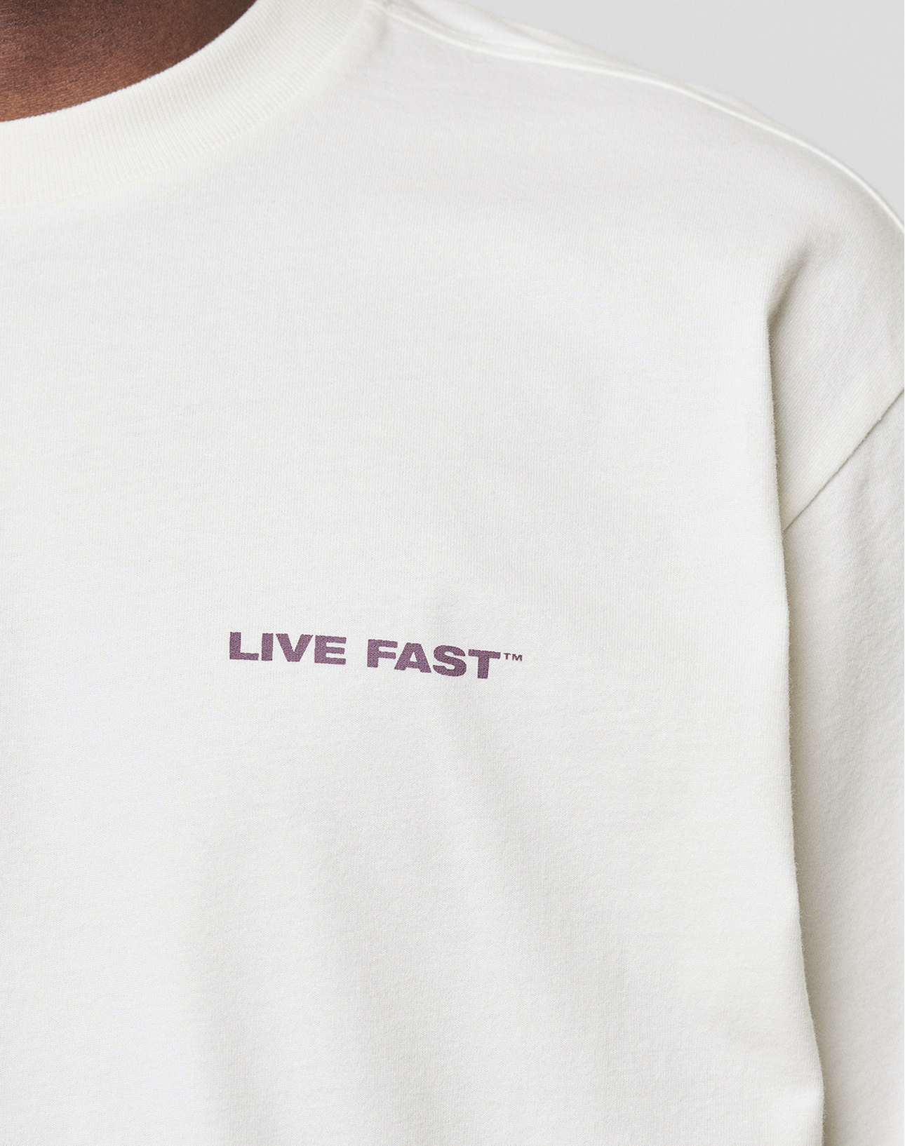 A close-up of the LFDY Grand Theft Tee by LIVE FAST DIE YOUNG, a white 100% Baumwolle crewneck with "LIVE FAST™" in small bold purple letters on the left chest.