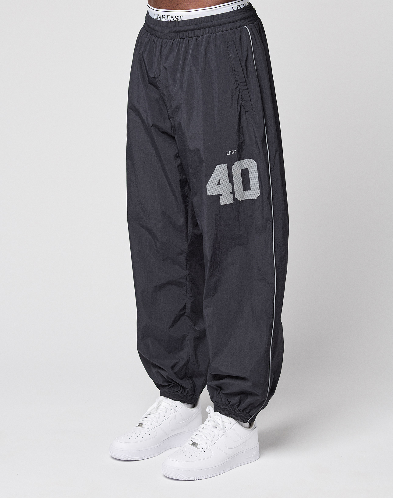 A person wears LIVE FAST DIE YOUNG LFDY Performance Trackpants in black, made of 100% nylon with a grey LIVE 40 reflektierender print on the left thigh, paired with white sneakers. Only the lower body is shown against a white background. | model