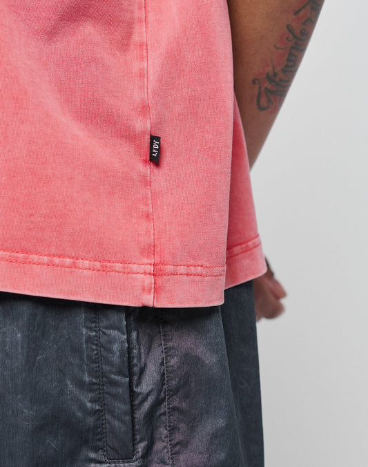 Close-up of a person in a pink Basic Evidence Tee by LIVE FAST DIE YOUNG, featuring a small black flag-label near the hem, paired with dark gray shorts. The tattooed arm is partially visible in the background.