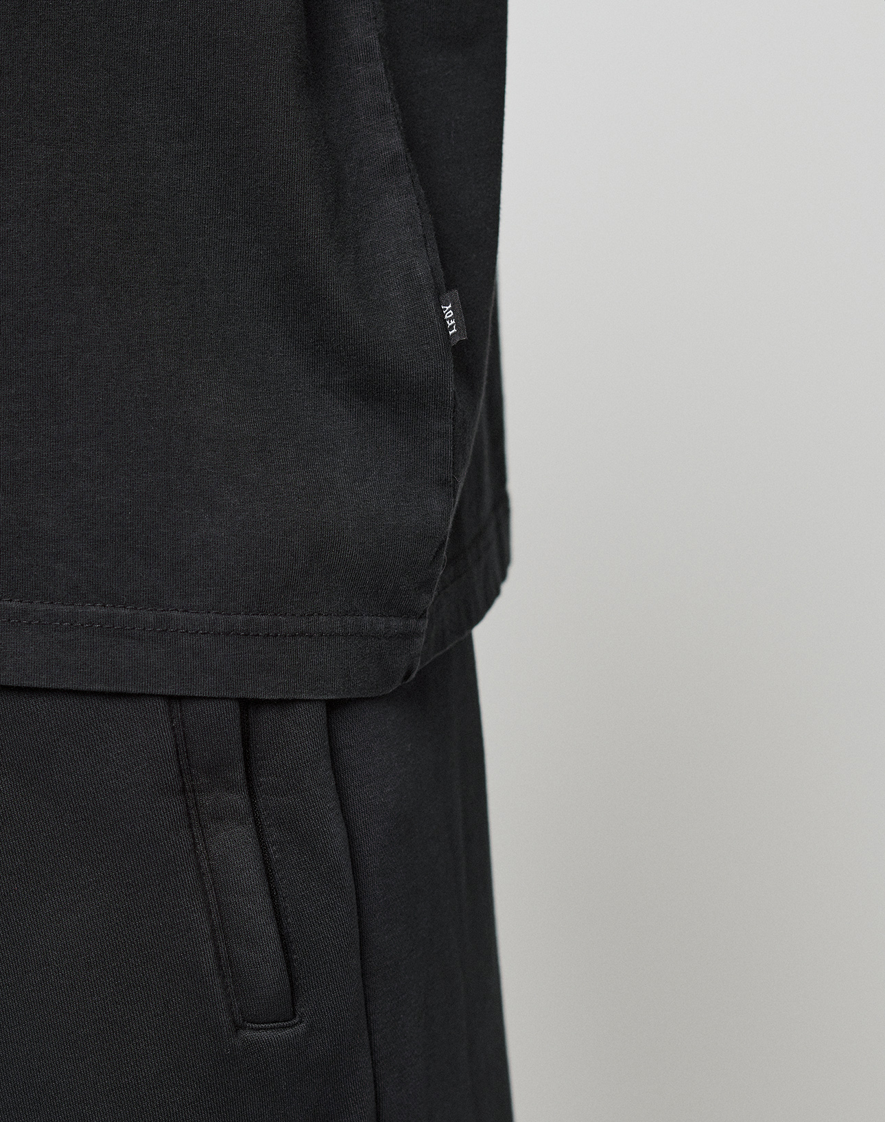 Close-up of the lower half of a black Oversize Fit Basic Evidence Tee by LIVE FAST DIE YOUNG, made from 100% Baumwolle, with a small side tag, paired with matching black pants featuring a visible pocket against a light background.