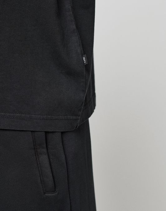 Close-up of the lower half of a black Oversize Fit Basic Evidence Tee by LIVE FAST DIE YOUNG, made from 100% Baumwolle, with a small side tag, paired with matching black pants featuring a visible pocket against a light background.