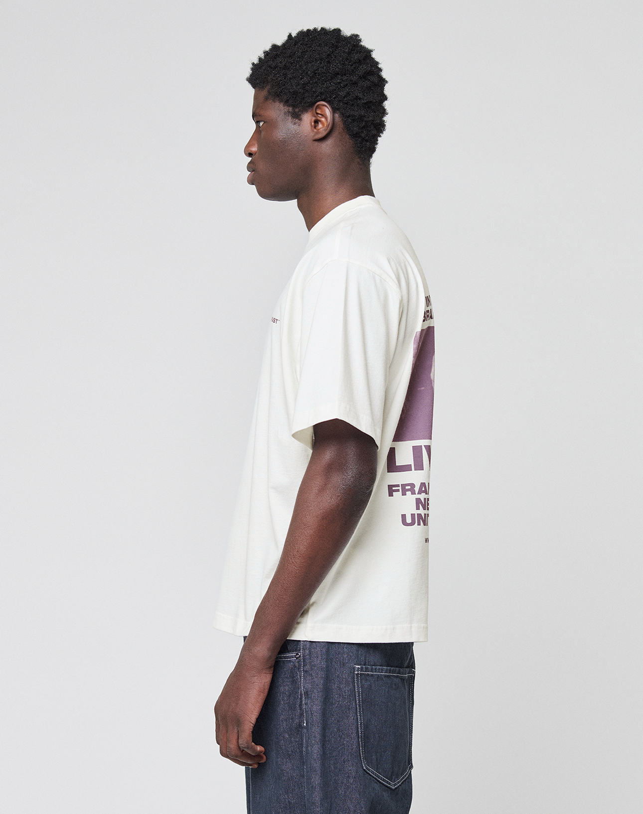 A person with short curly hair, wearing the LIVE FAST DIE YOUNG LFDY Grand Theft Tee—an oversized white 100% Baumwolle T-shirt with back text and graphics—and dark jeans stands in profile against a plain light gray background.